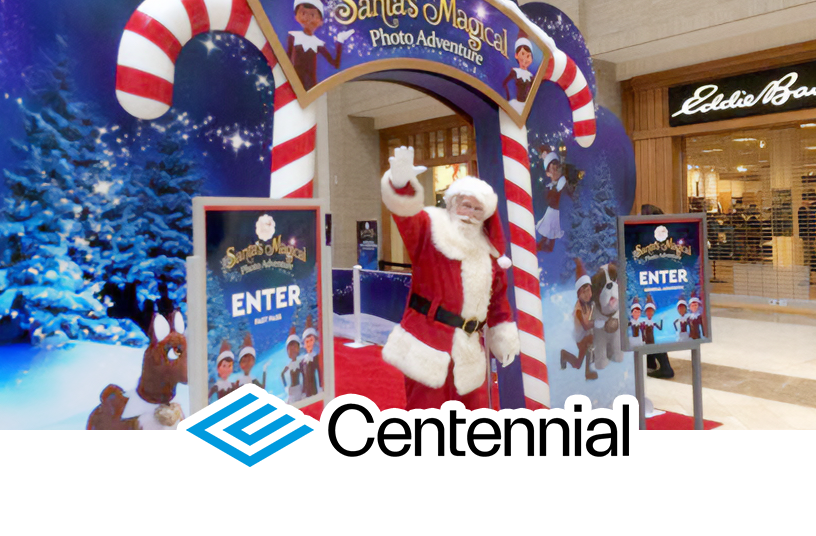 Santa Claus waving and standing underneath an arch made of two giant candy canes that says Santa's Magical Photo Adventure along the top. On the sides of the arch are signs that say Enter and feature the The Elf on the Shelf logo and Scout Elves along the bottom. On each side of the arch are Santaverse characters on a background with snow, pine trees, and magic sparkle trails. The Centennial logo overlays the bottom center of the photo.