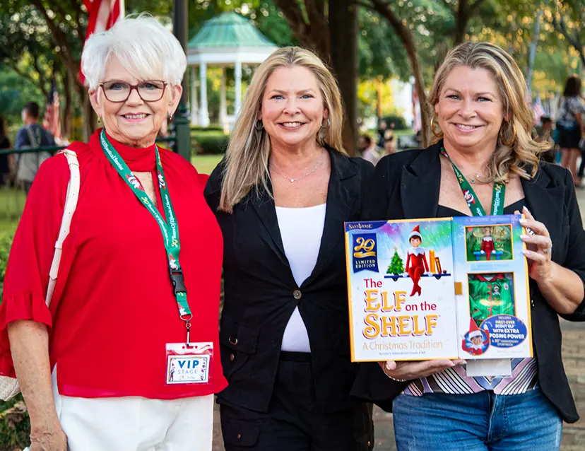 Carol Aebersold, Christa Pitts, and Chanda Bell standing in Marietta Square during the company's 20th anniversary celebration. Chanda is holding limited edition 20th anniversary The Elf on the Shelf boxset.