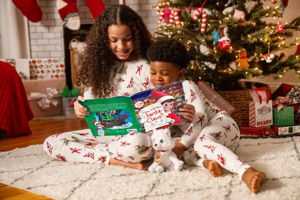 A young girl with black curly hair sits on the floor reading How Old is Santa Claus by Chanda A. Bell to her younger brother