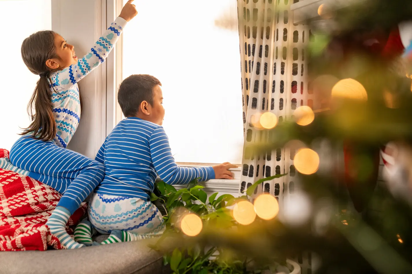A young boy and girl, in a living room decorated for Christmas, peer out a window as if searching for Santa Claus