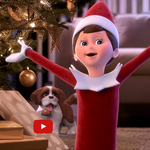 A snippet from the "Scout Elves Don't Rest" music video on YouTube, showing the boy Scout Elf singing in front of a wrapped Christmas present and decorated Christmas tree. Also in the background is an Elf Pets Saint Bernard pup. The YouTube play button is also featured on the image.