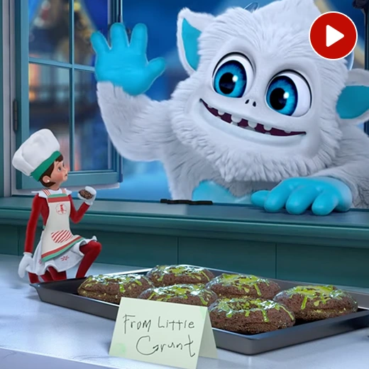 The SnoBiggie Yeti Little Grunt is leaning in through a window, waving hello to a girl elf who is wearing an apron and baker's hat. She is enjoying a batch of Little Grunt's iconic donuts he left for them on a tray on the counter. There is also a note reading, "From Little Grunt". In the top right corner is a video icon.