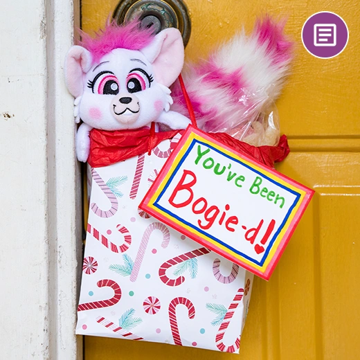 A pink and white Bogie plush peeking out of a candy cane patterned Christmas gift bag handing on a doorknob. Hanging off of the gift bag is a hand written sign that says "You've Been Bogie-d!". In the top right corner is a blog post icon.