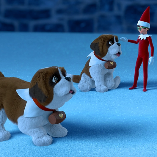 Two St. Bernard pups on a patch of snow with a stone retaining wall in the background. One pup is standing and the other is sitting down. A boy Scout Elf is standing next to the pup that is sitting down and has his arm raised up by his head and is about to pet him.