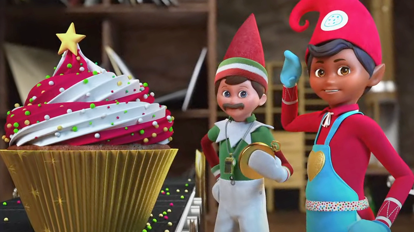 An Elf Mates® Chef is posed with Gym Dandy, who is holding a stop watch. The Elf Mates Chef is in going through the rigorous program of Top Elf Academy to learn his baking skillset. They are also posing with a giant cupcake with white and red frosting, topped with sprinkles and a golden star.
