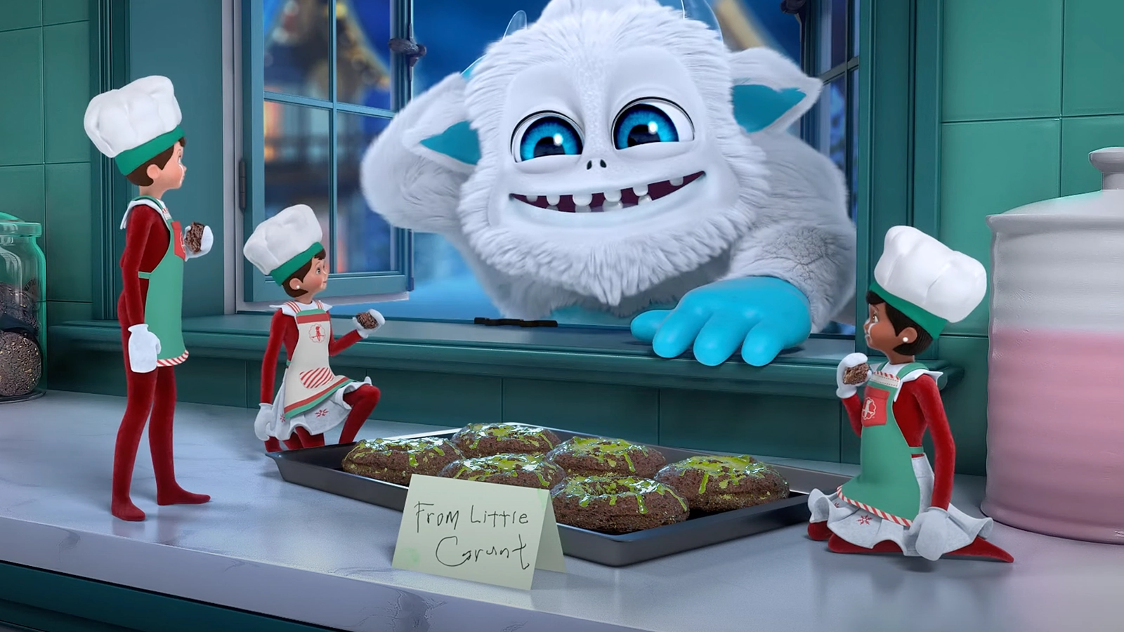 The SnoBiggie Yeti Little Grunt is leaning in through a window, interacting with a trio of Scout Elves who are all clad in aprons and baker's hats. They are enjoying a batch of Little Grunt's iconic donuts he left for them on a tray on the counter. There is also a note reading, "From Little Grunt".