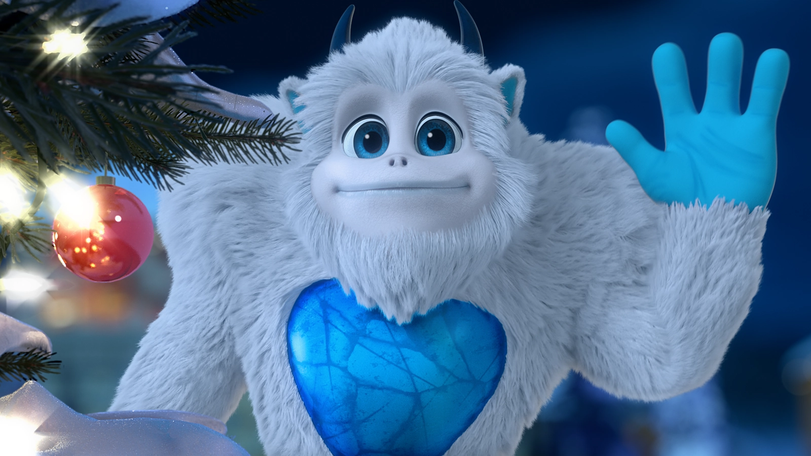 A SnoBiggie™ Yeti is holding his left hand up in a wave at the viewer. He is positioned partly behind a decorated Christmas tree.