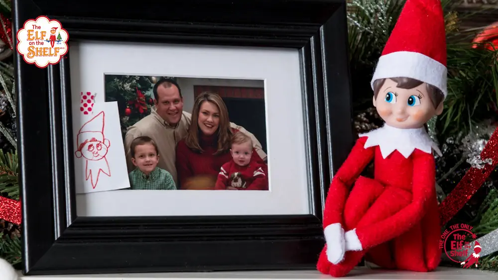 Elf on the Shelf scout elf facing a picture of a family with christmas decoration behind him.