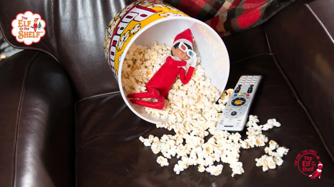 Elf on the Shelf in a box of popcorn on a chair with a remote.