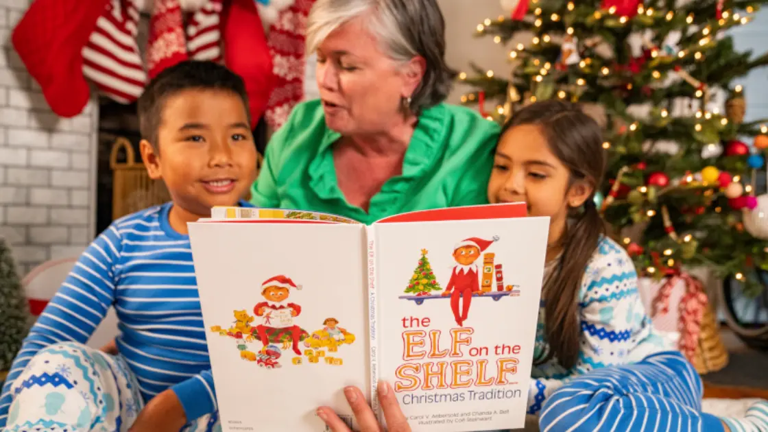Grandma with two children reading elf on the shelf book in front of christmas tree.webp