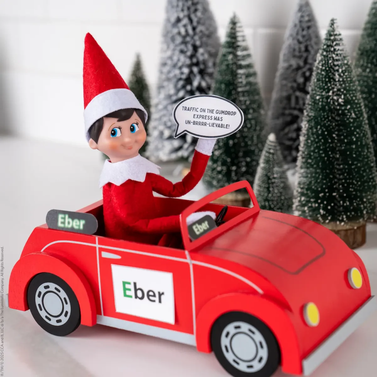 The Elf on the Shelf Scout Elf drives a red car with a sign on it that says Eber and holds a caption bubble that says, "Traffic on the Gumpdrop Express was unbelievable."