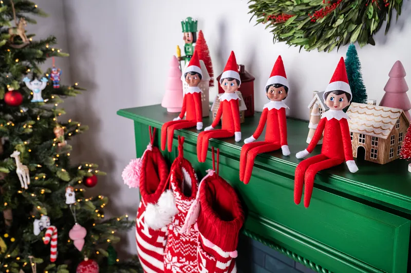 Four scout elves sitting on a green mantle with Christmas tree next to them