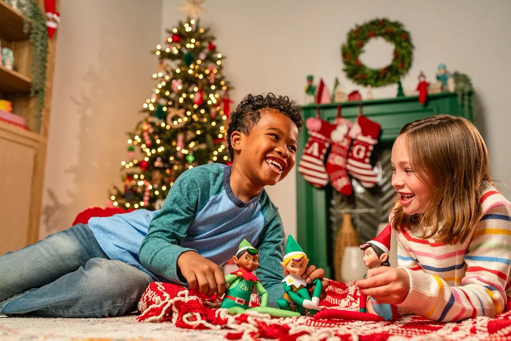 Children playing with Elf Mates dolls on a living room floor with Christmas tree and festive decorations in the background