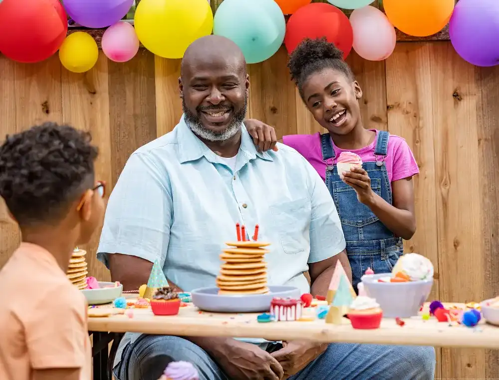 Father celebrating birthday with children outdoors, smiling dad with pancakes and candles while kids hold cupcakes at a colourful family party