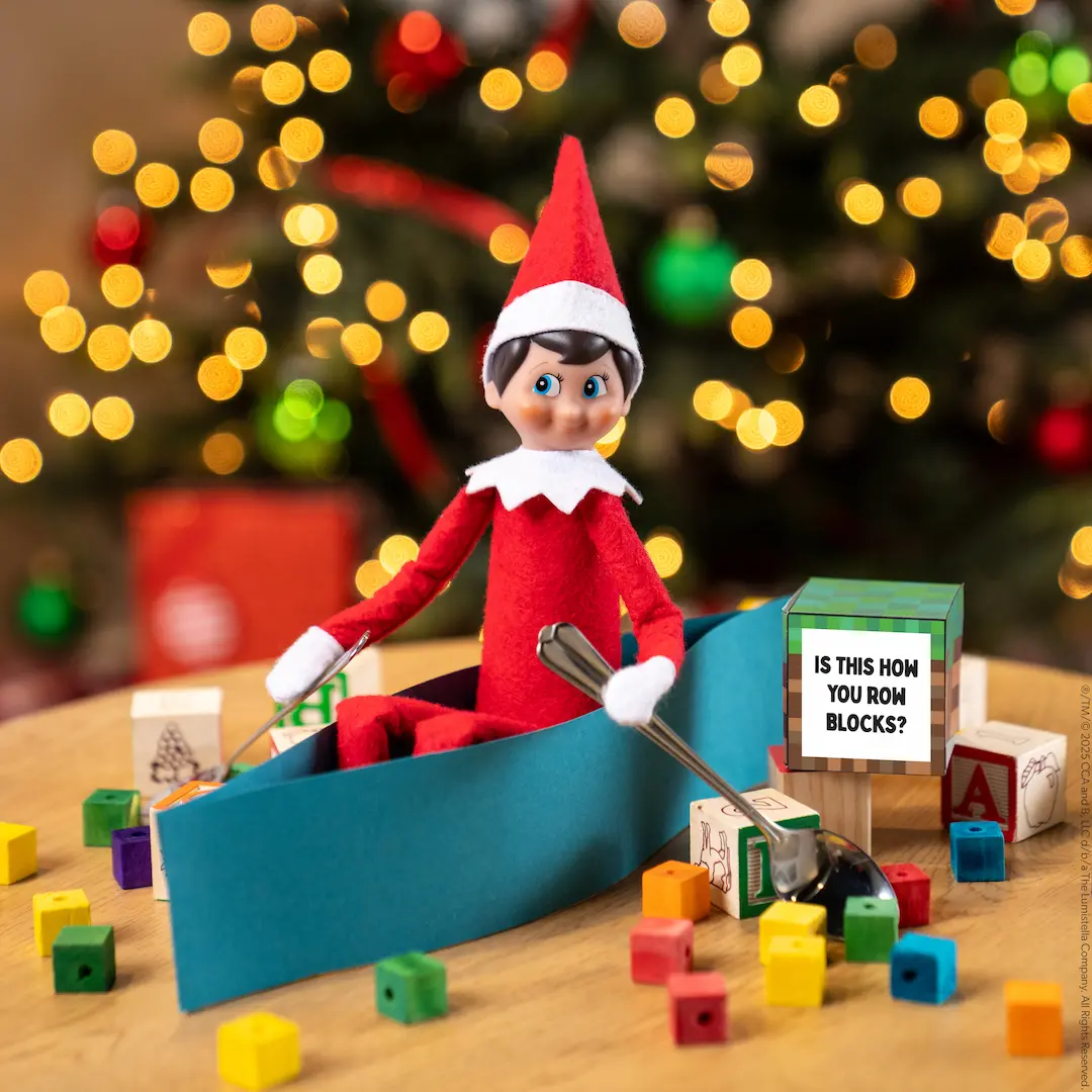 A Scout Elf rows a paper canoe using two spoons among scattered toy building blocks.