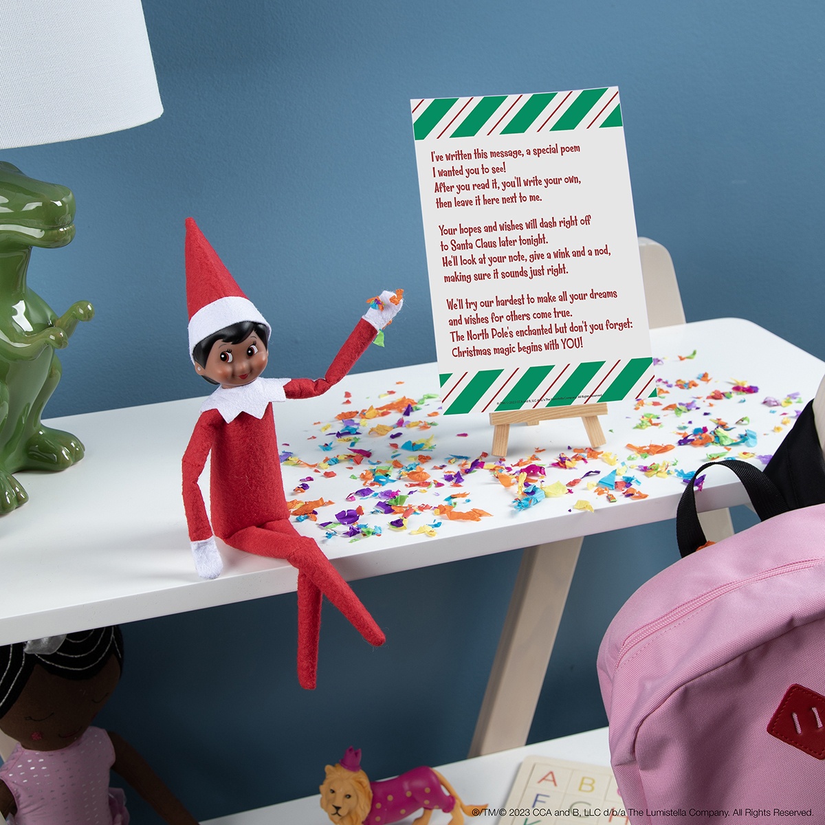 A Scout Elf sits on a desk beside a printed poem on a small easel, surrounded by colourful confetti.