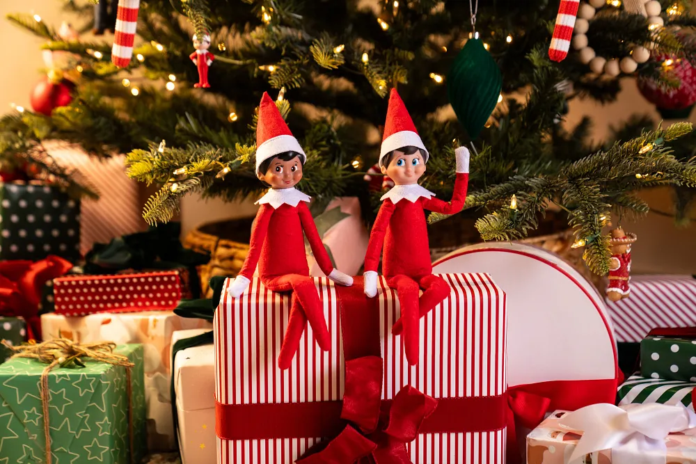Two Scout Elves sitting on wrapped Christmas presents beside a decorated Christmas tree