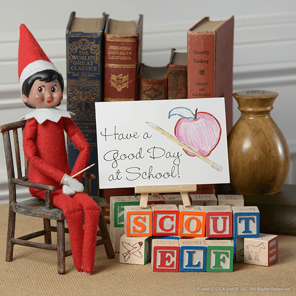 A Scout Elf sitting on a small wooden chair beside a Have a Good Day at School sign, alphabet blocks spelling Scout Elf and a row of books