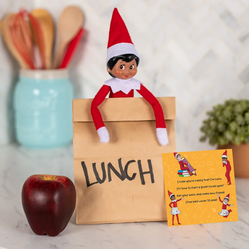 A Scout Elf peeking out of a paper lunch bag labelled Lunch, with a red apple and a handwritten encouraging note