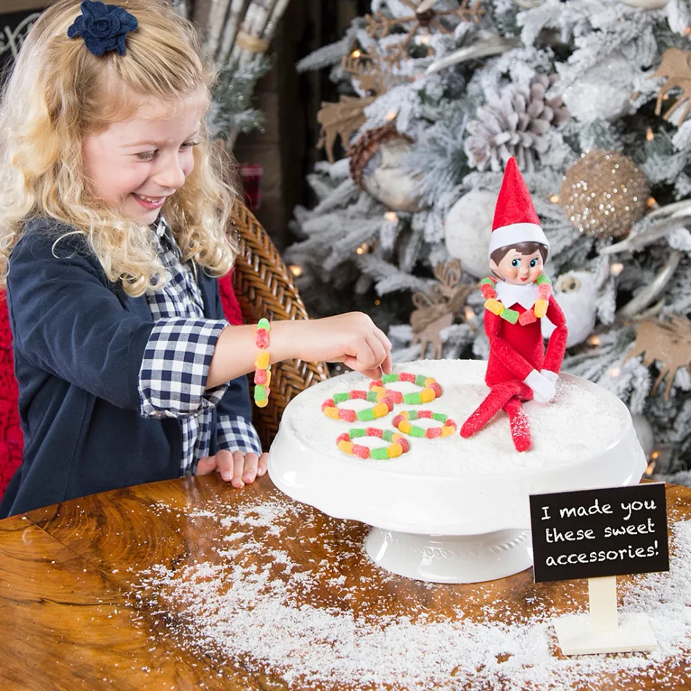 A smiling girl discovering a Scout Elf beside a cake stand of sweets, with a sign reading I made you some sweet accessories