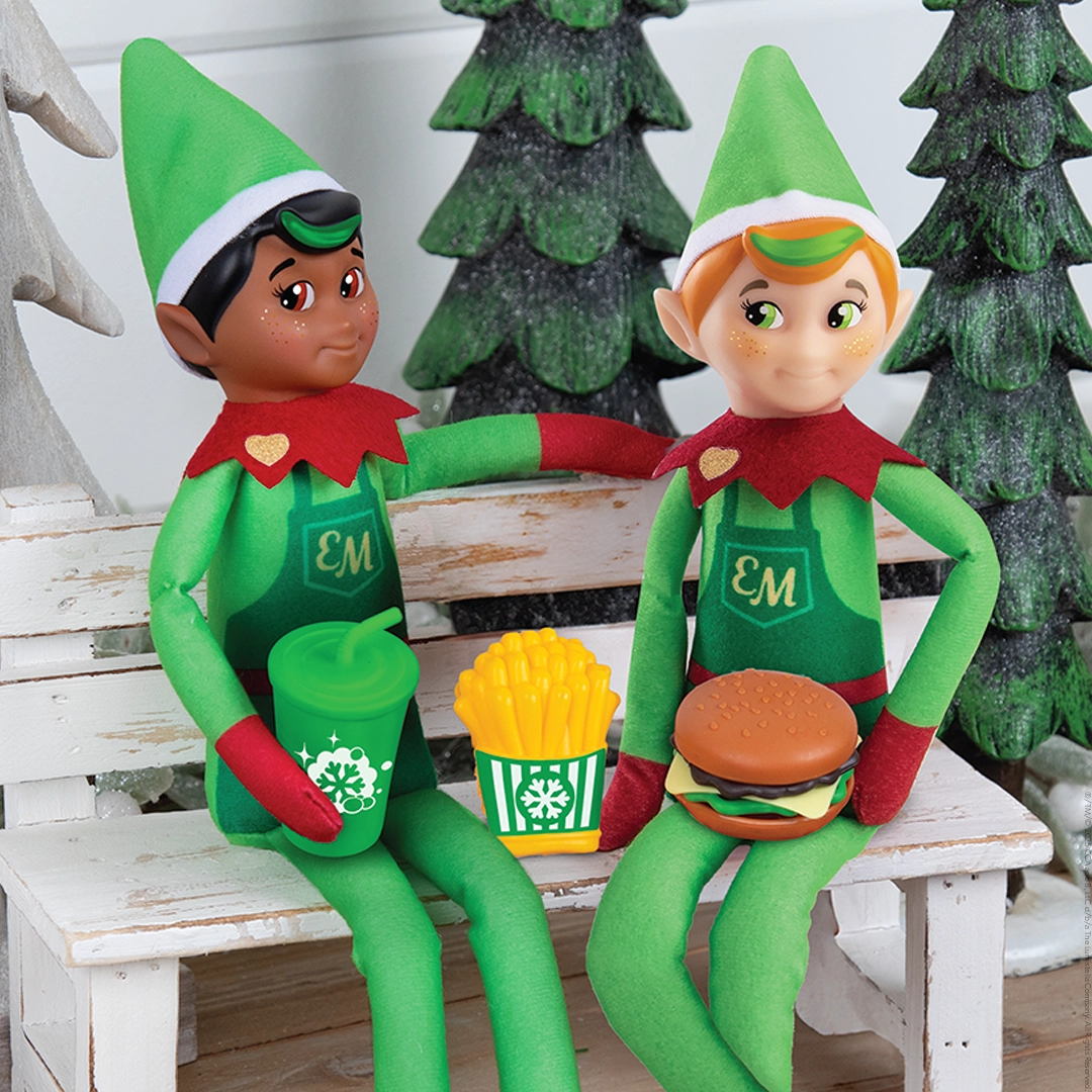A pair of Elf Mates® Cobblers are enjoying a hamburger, fries and soda while taking a break on a white elf-sized bench. Behind them, you can see a trio of decorative conifer trees.