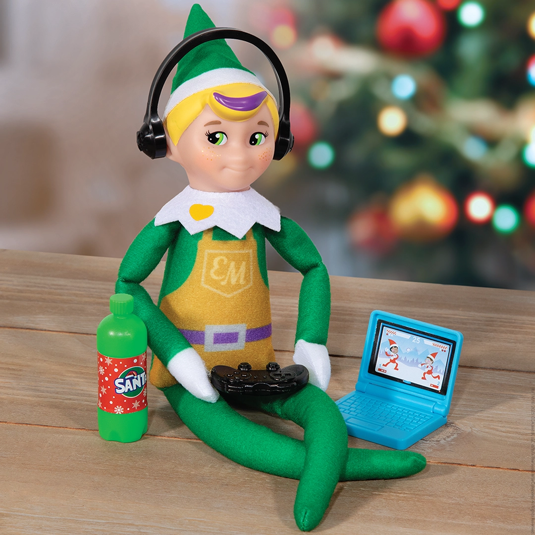 An Elf Mates® Toy Maker sits with a gaming laptop, a bottle of Santa Soda, and controller while wearing headphones. A blurry, festively decorated Christmas tree can be made out in the background.